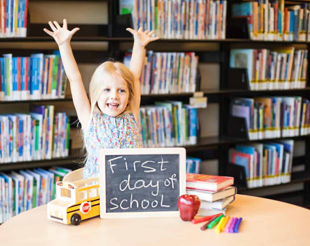 A girl stands in front of a library with her hands raised smiling a sign on a table in front of her says first day of school