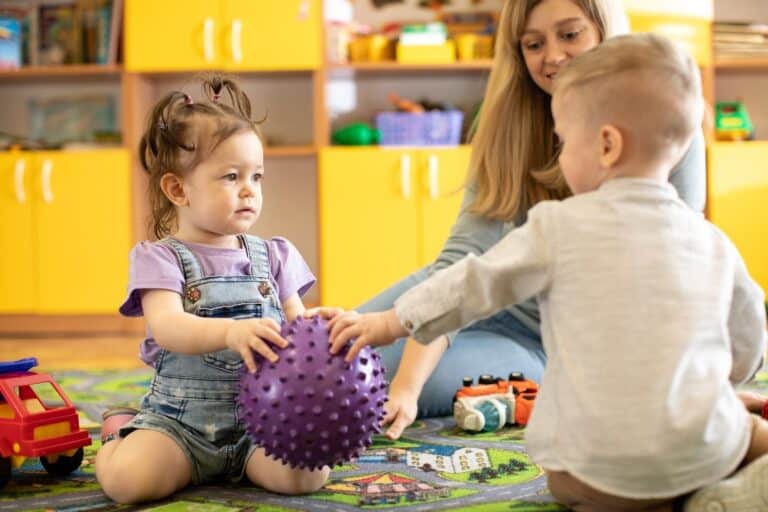 A girl holding a toy while a boy reaches for it. A teacher sits behind them