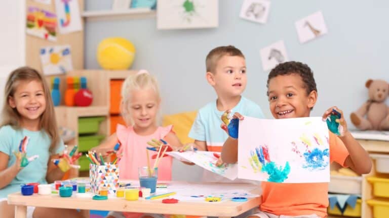 An art classroom three children sit at a table a 4th child holds a picture up to the camera