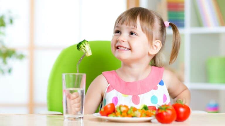 A girl smiles holding a fork with a piece of broccoli on it in front of her sits a plate of vegetables with some tomatoes on the side