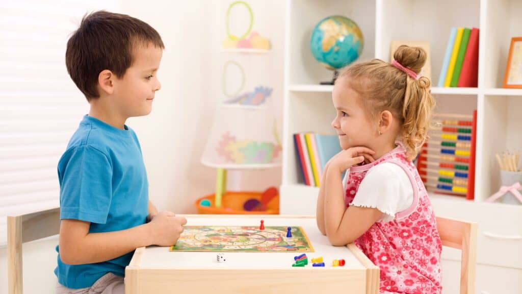 An elementary school boy and girl sit on opposite sides of a child table playing a board game