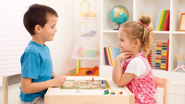 An elementary school boy and girl sit on opposite sides of a child table playing a board game