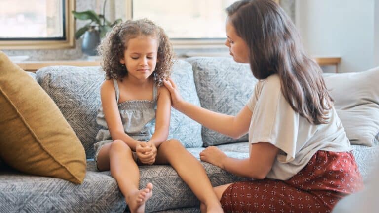 An upset girl sits on a couch an older girl comforts her