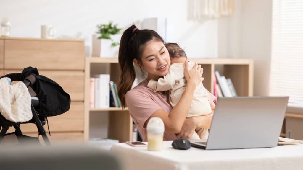 A woman burps her baby while looking at a laptop