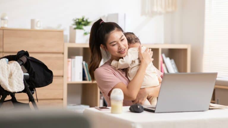 A woman burps her baby while looking at a laptop