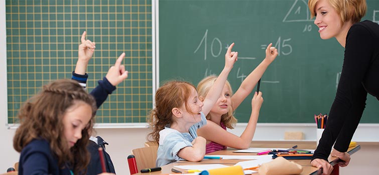 Children sitting at desks raising their hands while a teacher smiles at them