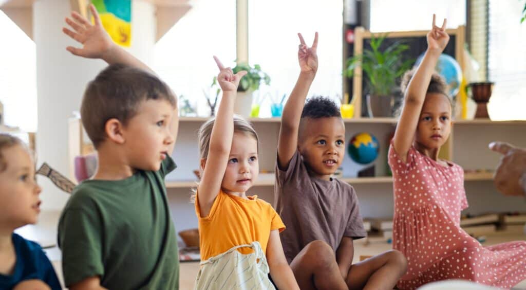 Five children sit on the ground in an elementary school classroom holding their hands up showing two fingers