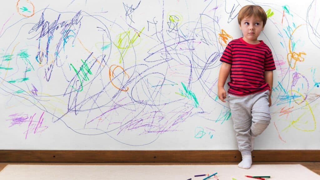 A chagrined boy stands against a white wall covered in crayon marks