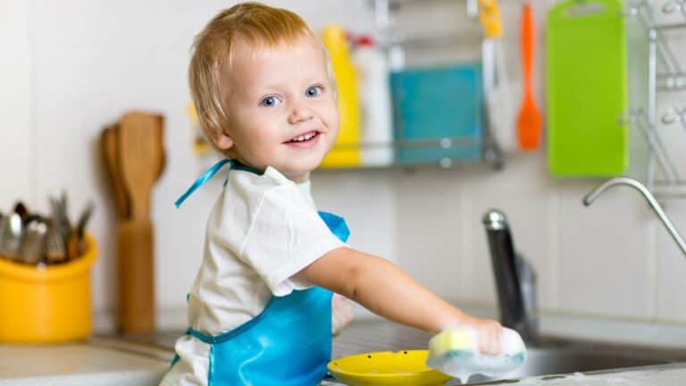 a young boy washes dishes and smiles at the camera
