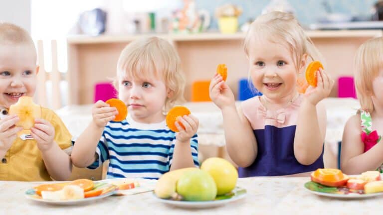 Children sitting at a table holding orange slices With plates of fruit in front of them